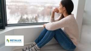 Ventanas Retalux, ventanas de PVC y aluminio en Madrid, Una mujer con jersey y vaqueros está sentada junto a una ventana mirando hacia el exterior. El logotipo "RETALUX" aparece en la esquina inferior izquierda.