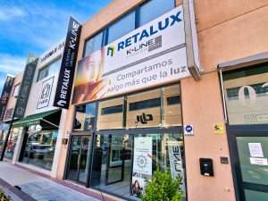 Ventanas Retalux, ventanas de PVC y aluminio en Madrid, Fachada de edificio comercial con tienda RETALUX con gran cartelería, flanqueada por otras tiendas con diferentes carteles bajo un cielo azul. Con elegantes Ventanas de Aluminio K-Line, la estructura irradia modernidad y estilo.