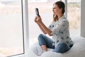 Ventanas Retalux, ventanas de PVC y aluminio en Madrid, Mujer sentada en un sillón puf blanco junto a una ventana con aislamiento acústico ventanas, sosteniendo una tableta, mirando la pantalla, con un paisaje urbano visible afuera.
