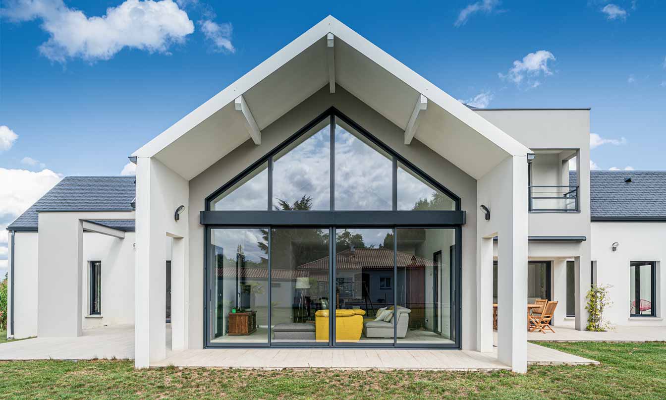 Ventanas Retalux, ventanas de PVC y aluminio en Madrid, Casa moderna con grandes ventanales, techo a dos aguas y césped bajo un cielo azul con nubes dispersas.