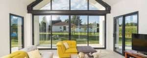 Ventanas Retalux, ventanas de PVC y aluminio en Madrid, Sala de estar luminosa con grandes puertas de vidrio K-Line, muebles amarillos y vista a un patio trasero verde. Los gatos descansan en el piso.