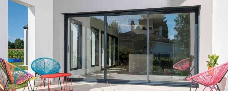 Ventanas Retalux, ventanas de PVC y aluminio en Madrid, Un patio moderno con sillas de colores y una gran puerta corrediza de vidrio que refleja un jardín y una casa.