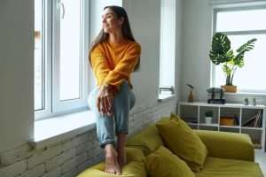 Beautiful young woman looking through a window and smiling while sitting on the window sill